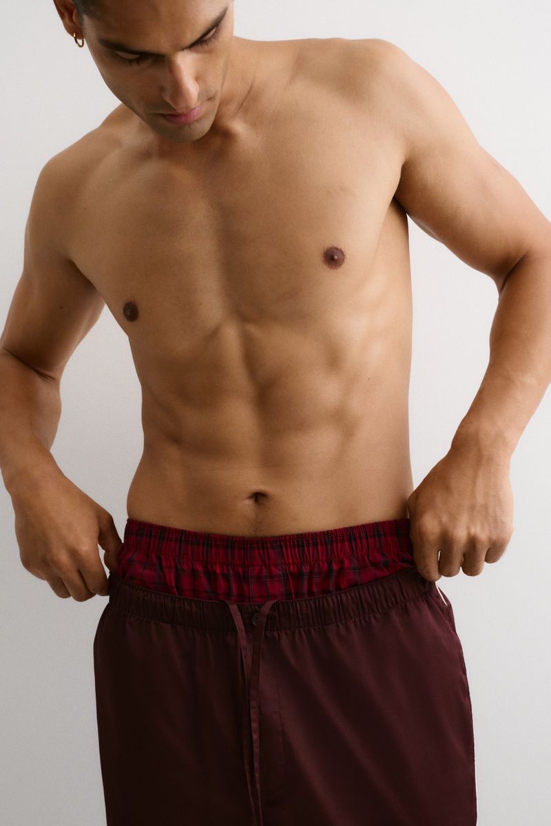 Shirtless athletic man adjusts maroon athletic shorts against a plain white background in a medium studio shot, abdomen in focus.