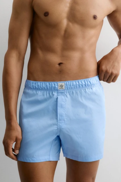Torso shot of a light-skinned adult male wearing light blue boxer shorts against an off-white background in a brightly lit photo studio.