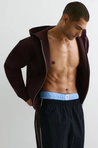 Man wearing an unzipped dark hoodie and dark pants with light blue boxer shorts against a plain white background
