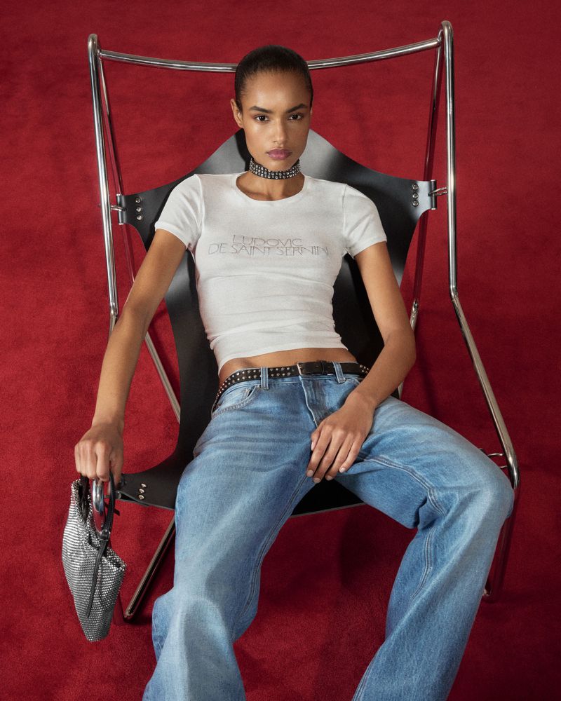 Model Ana Beatriz Cortes seated in a black sling chair wearing a white t-shirt and blue jeans over a red carpet.