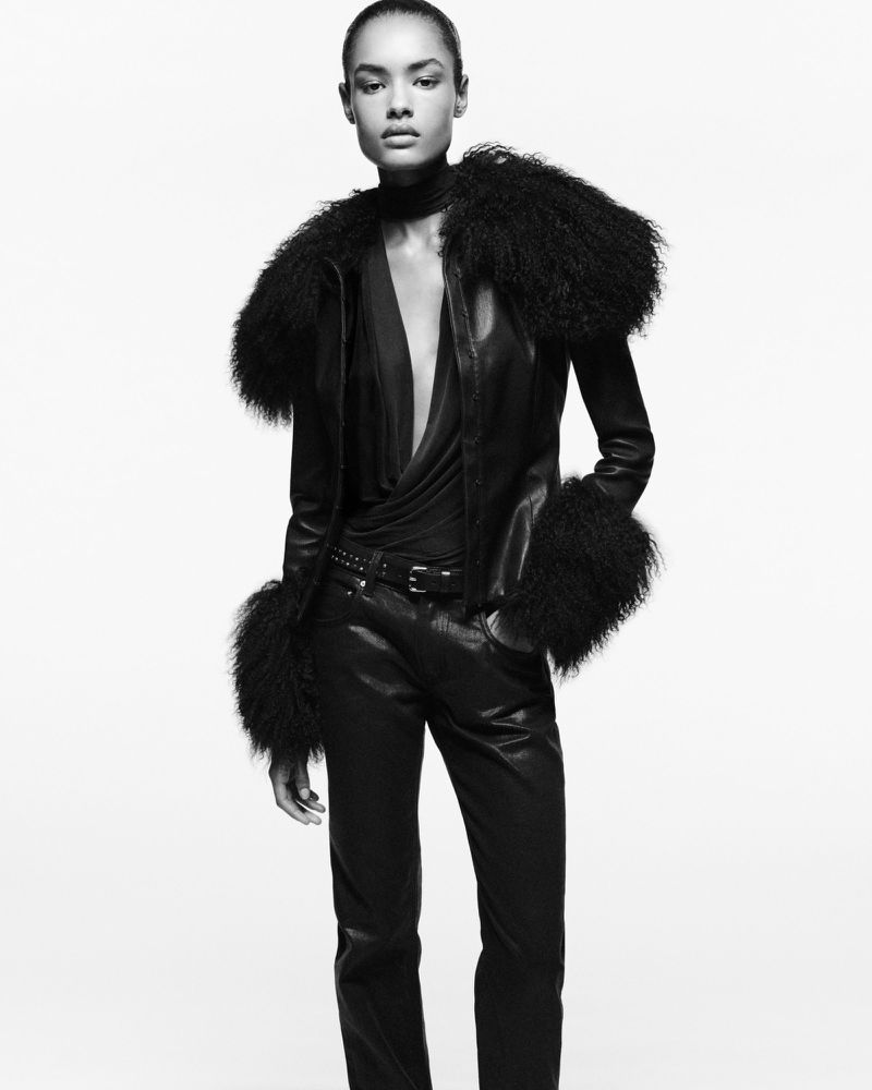 Elegant woman in black leather jacket and pants with dramatic fur accents posing in a fashion shot against a white backdrop.