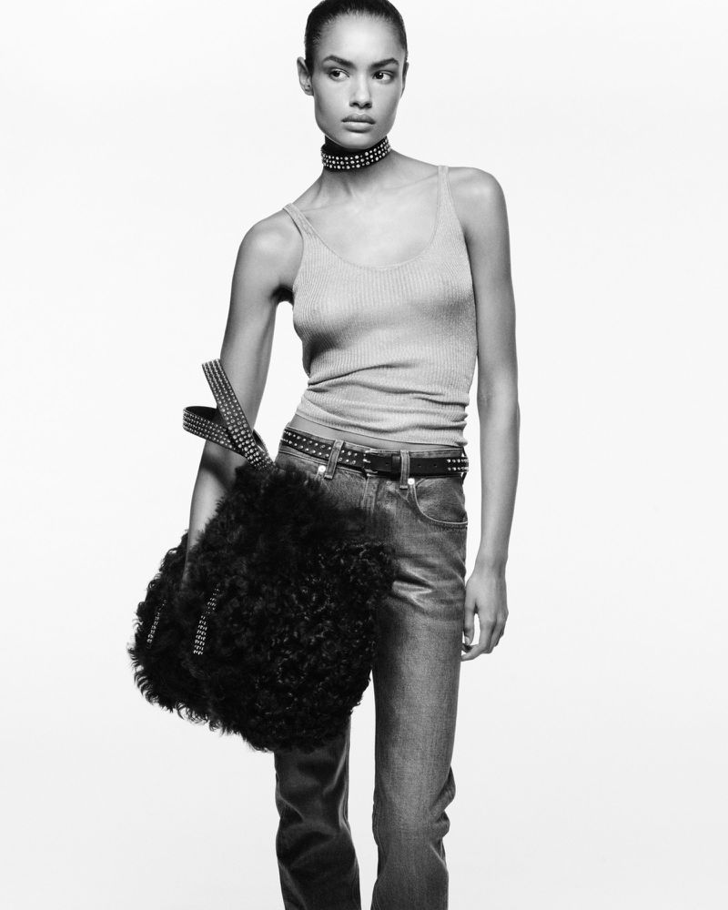 A model poses in a studio wearing jeans, a tank top, a studded belt and choker, and holding a furry black handbag in black and white.