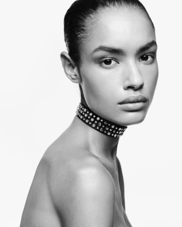 Monochrome close-up portrait of model Ana Beatriz Cortes wearing a studded choker necklace against a bright white background.