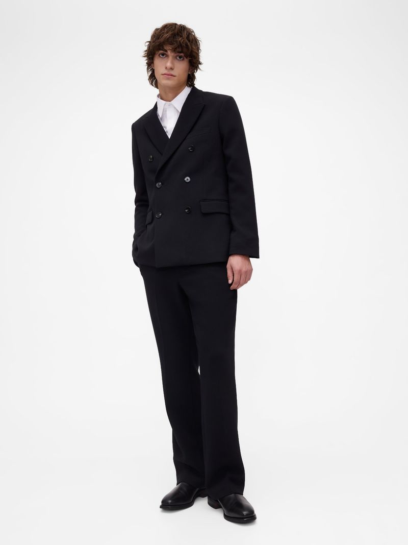 Adult male in a black double-breasted suit with a white collared shirt stands indoors against a white background in a studio shot.