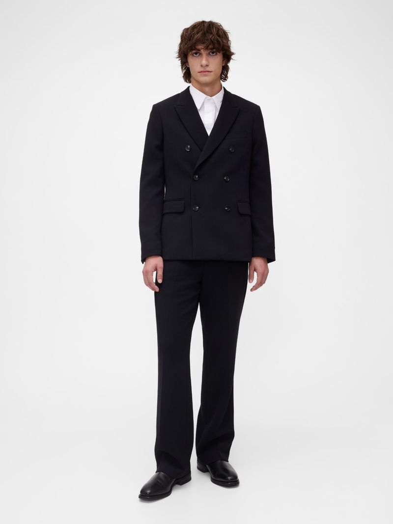 Full-length shot of a young adult man in a black double-breasted suit with flared trousers, paired with a white dress shirt, against a white backdrop.