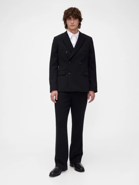 Full-length shot of a young adult man in a black double-breasted suit with flared trousers, paired with a white dress shirt, against a white backdrop.