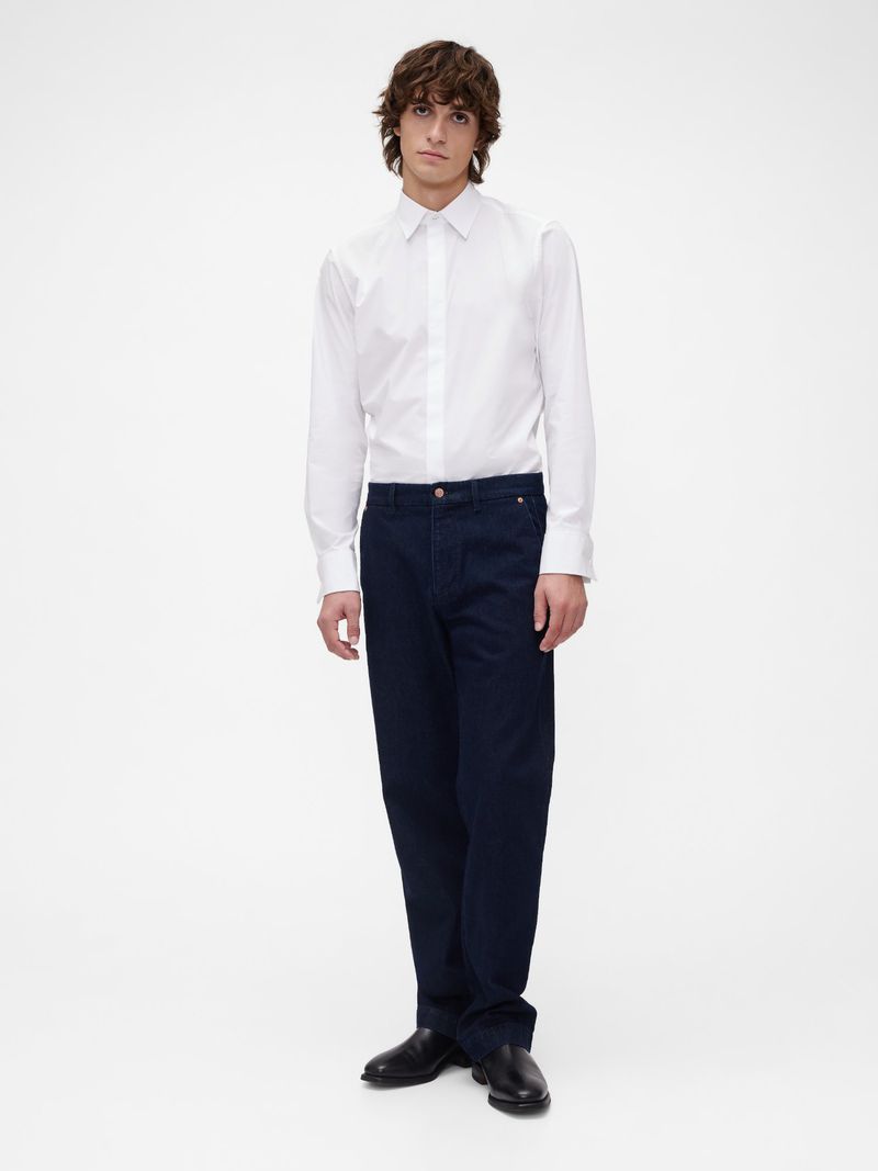 Full body portrait of a male model posing in a white collared long-sleeved shirt and dark blue jeans against a plain white backdrop.