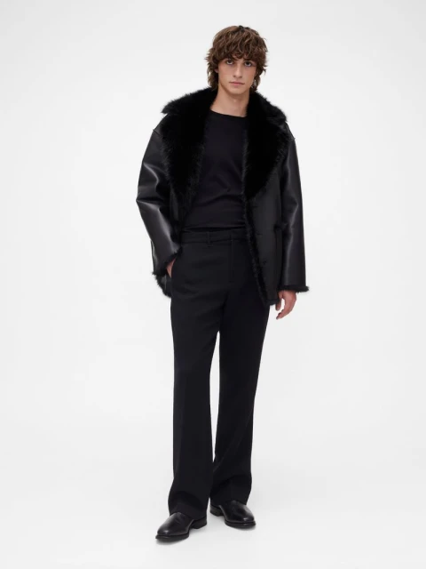Full-length shot of a man in a black shearling jacket, black shirt, and black trousers, posing against a white background.