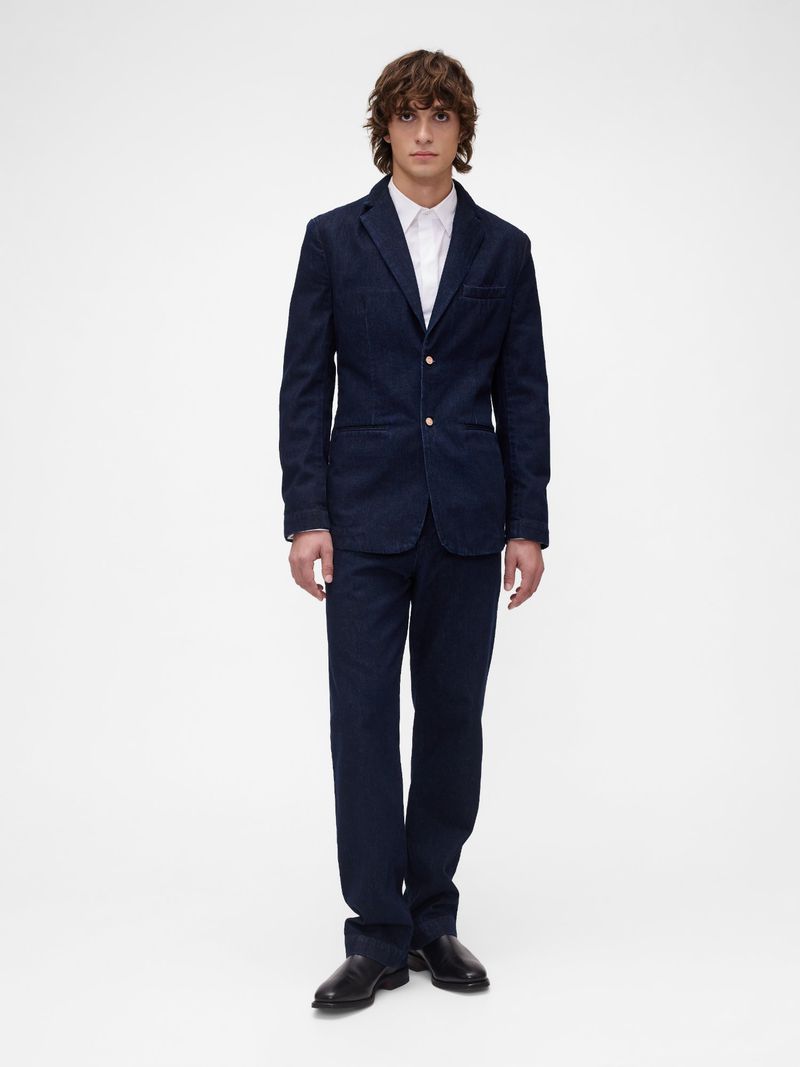Full-length shot of a man in a blue denim suit and a white dress shirt, standing against a bright white backdrop in a studio setting.