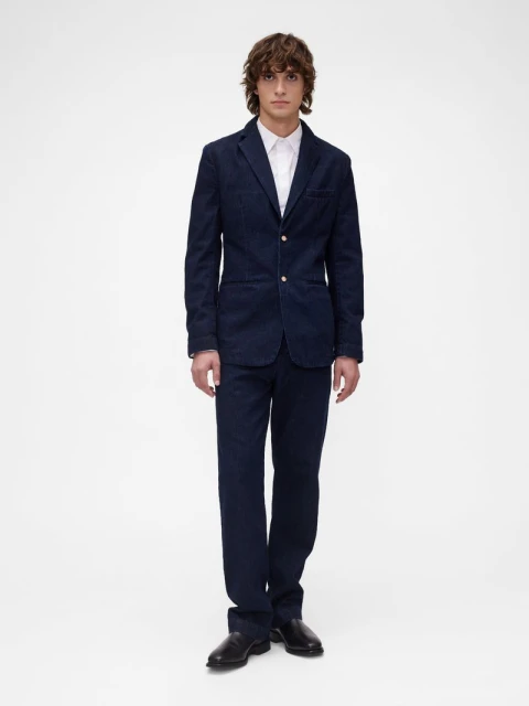 Full-length shot of a man in a blue denim suit and a white dress shirt, standing against a bright white backdrop in a studio setting.