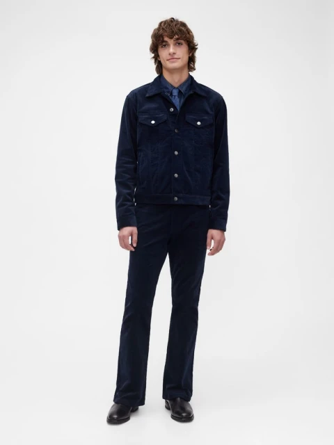 Full-body shot of a man in a navy corduroy jacket and matching pants. He is standing indoors in front of a white background.