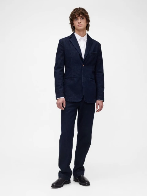 A young adult male stands in a studio wearing a navy blue suit with a white shirt and black leather shoes against a white backdrop.