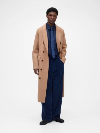 Model wearing a long camel double-breasted coat, blue button-down shirt, blue tie, and wide navy corduroy trousers on a white backdrop.