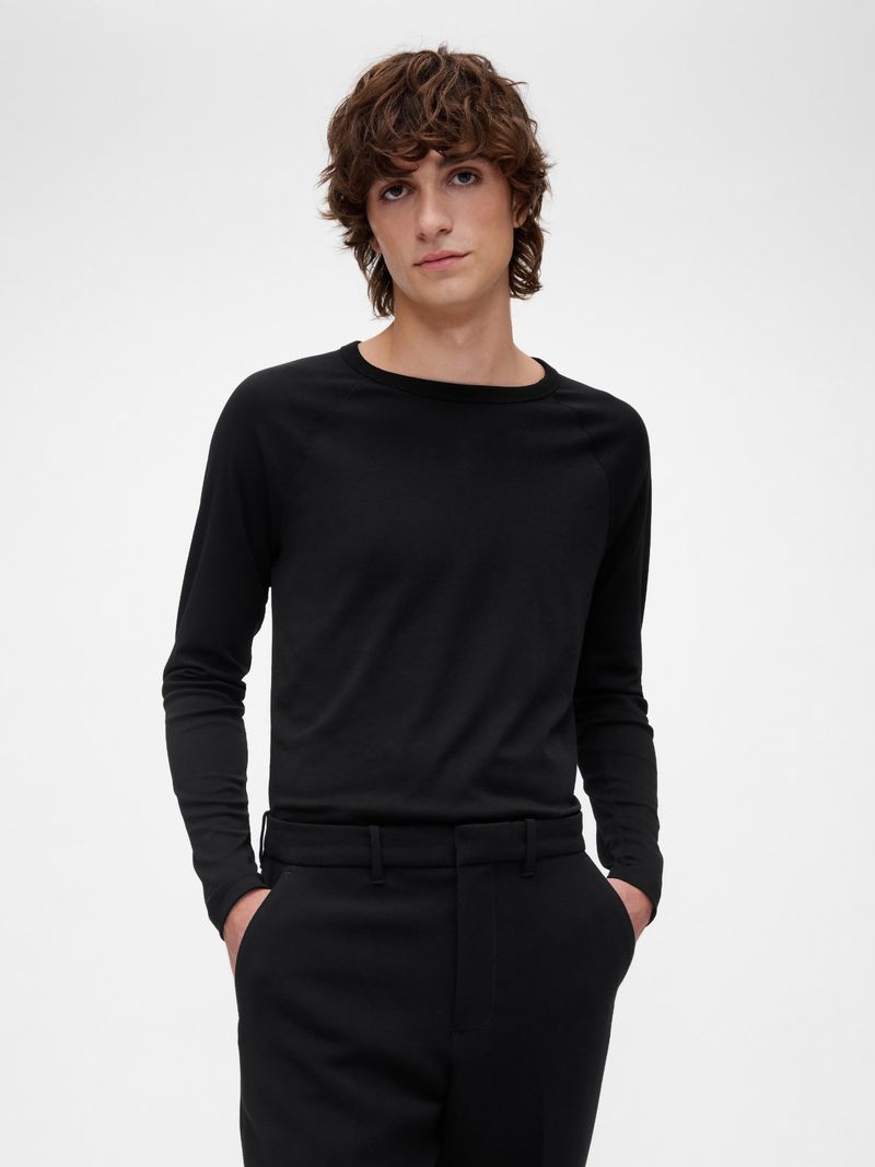 A young adult man poses indoors, wearing a long-sleeved black shirt and black pants, standing against a solid white backdrop in a portrait.