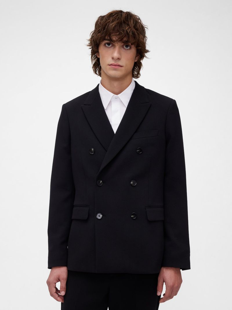 Man with wavy brown hair wearing a black double-breasted blazer and a white collared shirt, posing in front of a white background.