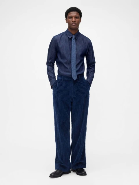 Full-length studio shot of an adult male model in a denim shirt, tie, and wide wale corduroy trousers against a plain white backdrop.