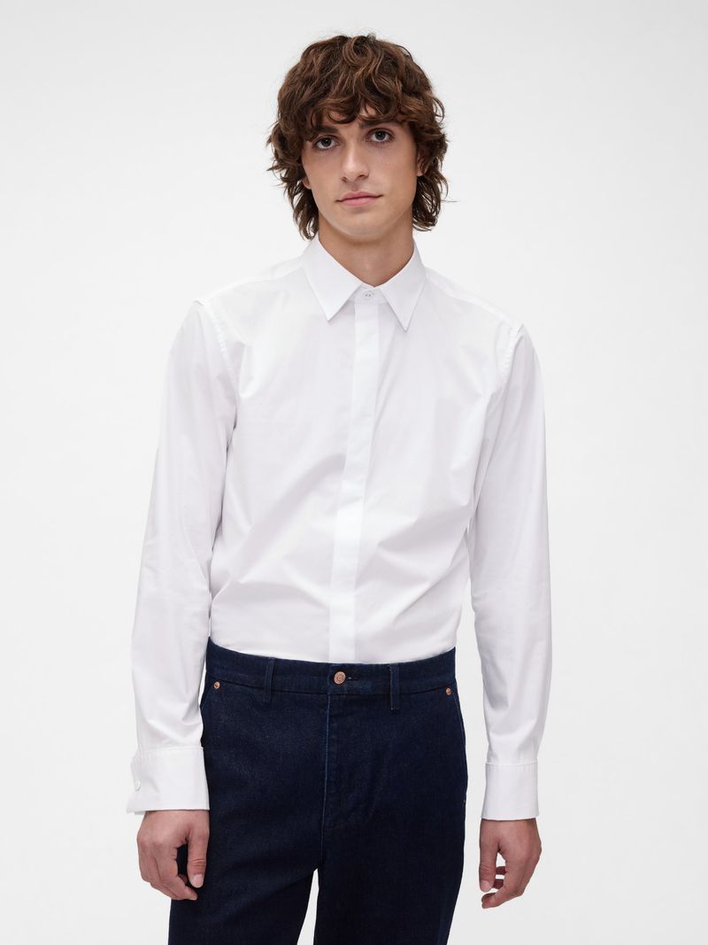 Close-up studio shot of a young adult male with wavy dark hair, wearing a white button-down shirt and dark jeans, against a white backdrop.
