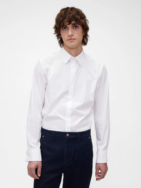 Close-up studio shot of a young adult male with wavy dark hair, wearing a white button-down shirt and dark jeans, against a white backdrop.