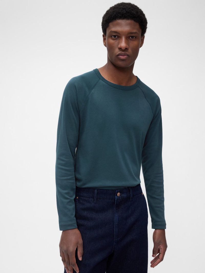 Studio shot of an adult male fashion model wearing a long-sleeve teal shirt and dark blue jeans against a plain white backdrop.