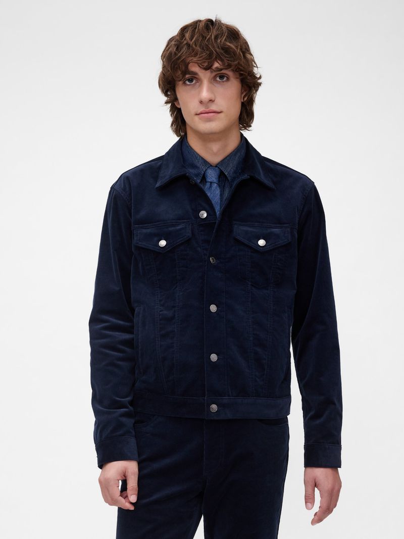 A young adult male stands in a photo studio wearing a navy corduroy jacket and matching pants against a neutral backdrop, fashion shot.