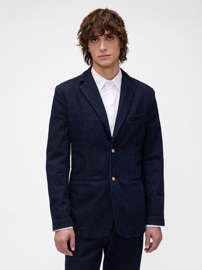 Studio shot of a male fashion model in a dark blue denim suit and white button-down shirt, posing against a bright white backdrop.