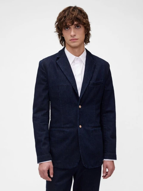 Studio shot of a male fashion model in a dark blue denim suit and white button-down shirt, posing against a bright white backdrop.