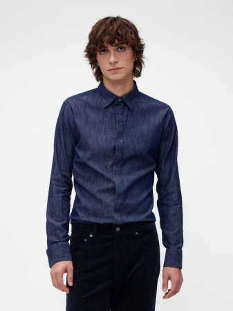 Model wearing a dark blue denim button-up shirt and dark trousers against a white seamless studio backdrop.