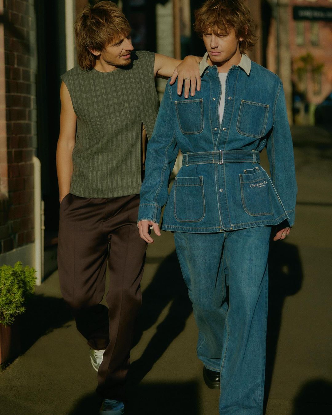 Two adult men pose outdoors wearing a denim jacket with a belt, a sleeveless top, and pants, standing in front of a brick building.
