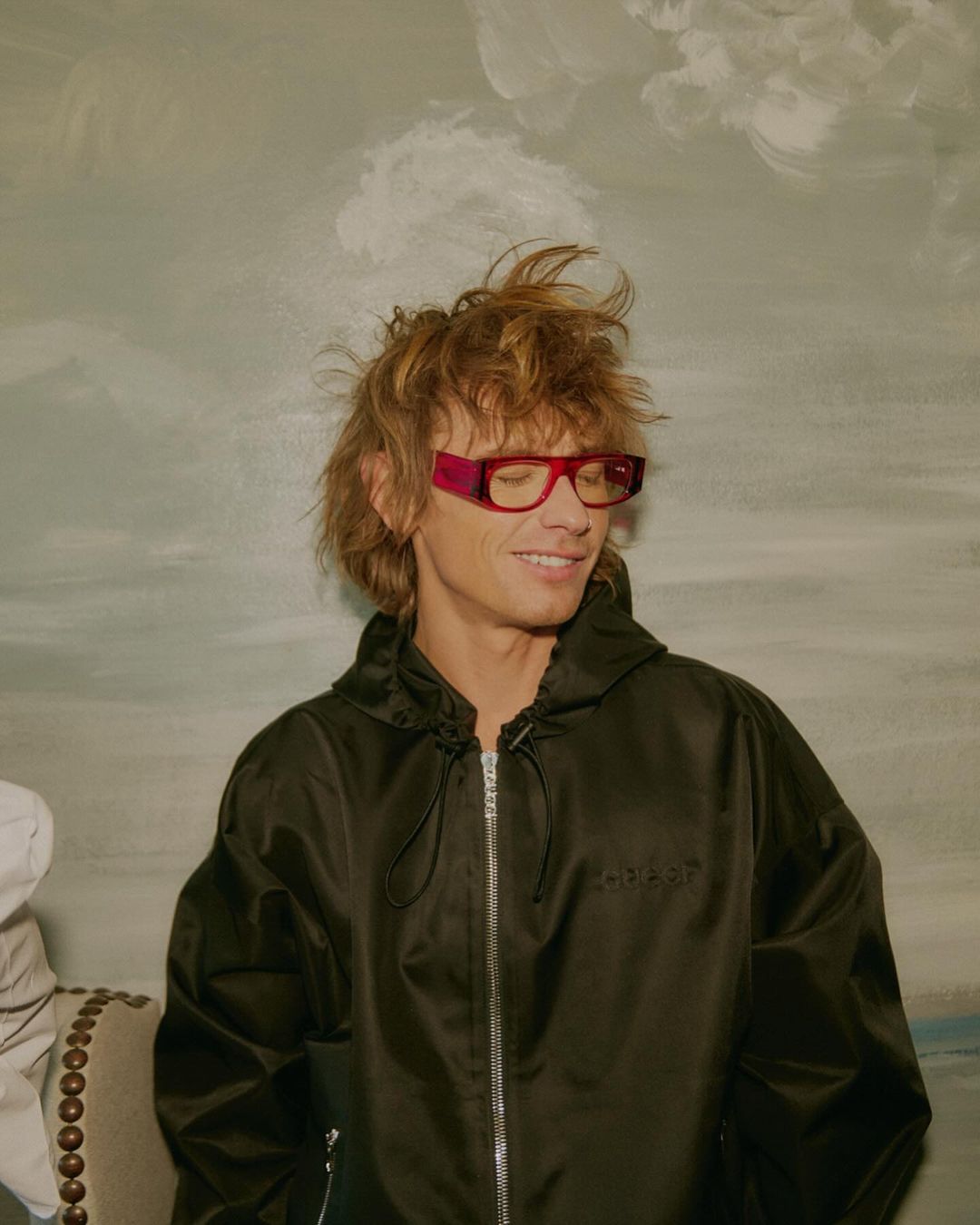 Smiling adult man with tousled hair wearing red glasses and black hooded jacket, posing indoors against a painted landscape backdrop.