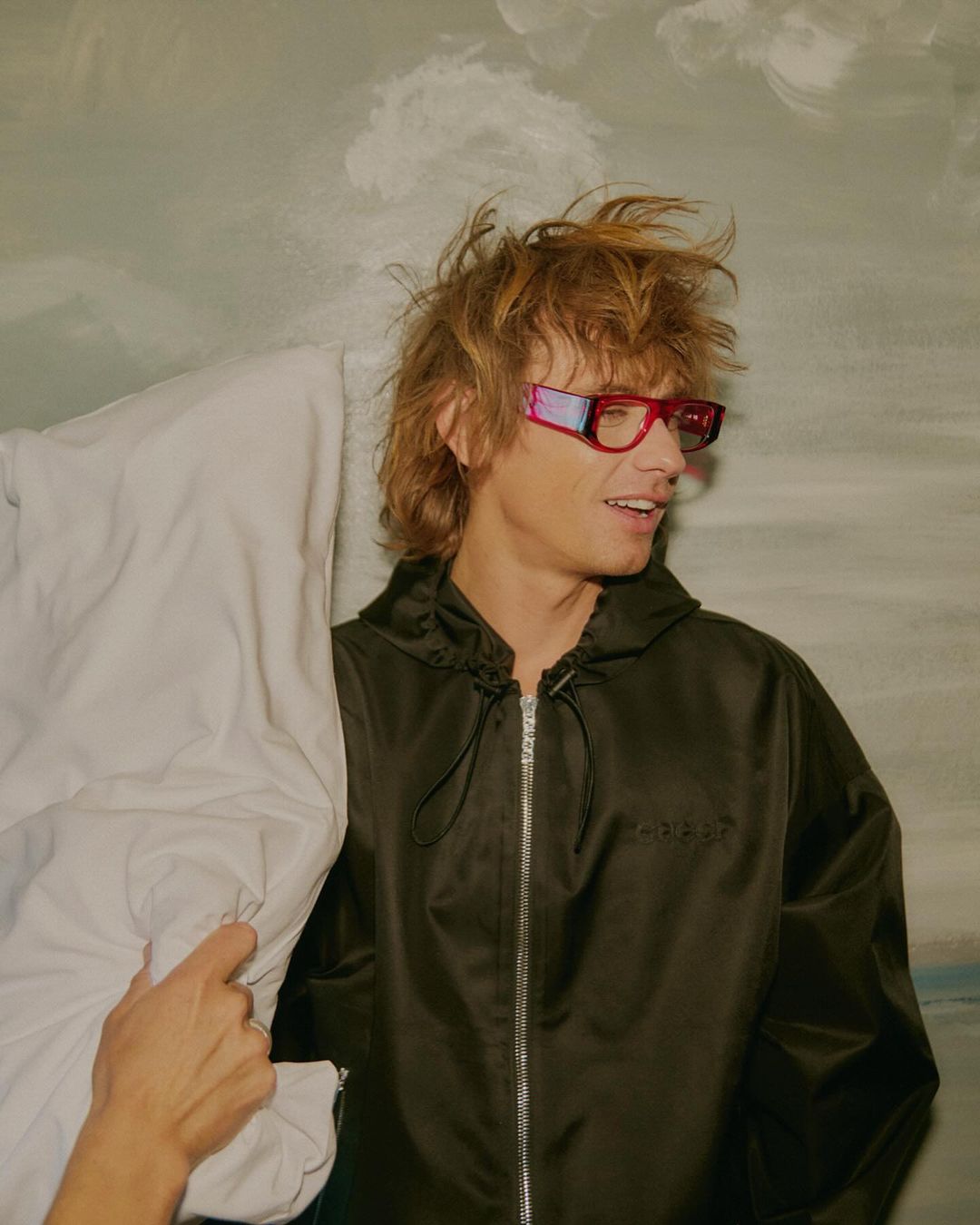 Man with tousled blond hair wearing a black jacket and pink sunglasses holding a white pillow against a painted background
