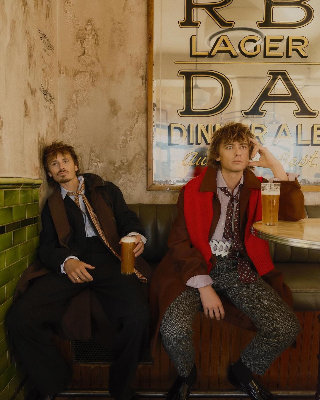 Two young adult men are sitting at a cafe table next to a wall with an advertisement for RB Lager, casually drinking beer.