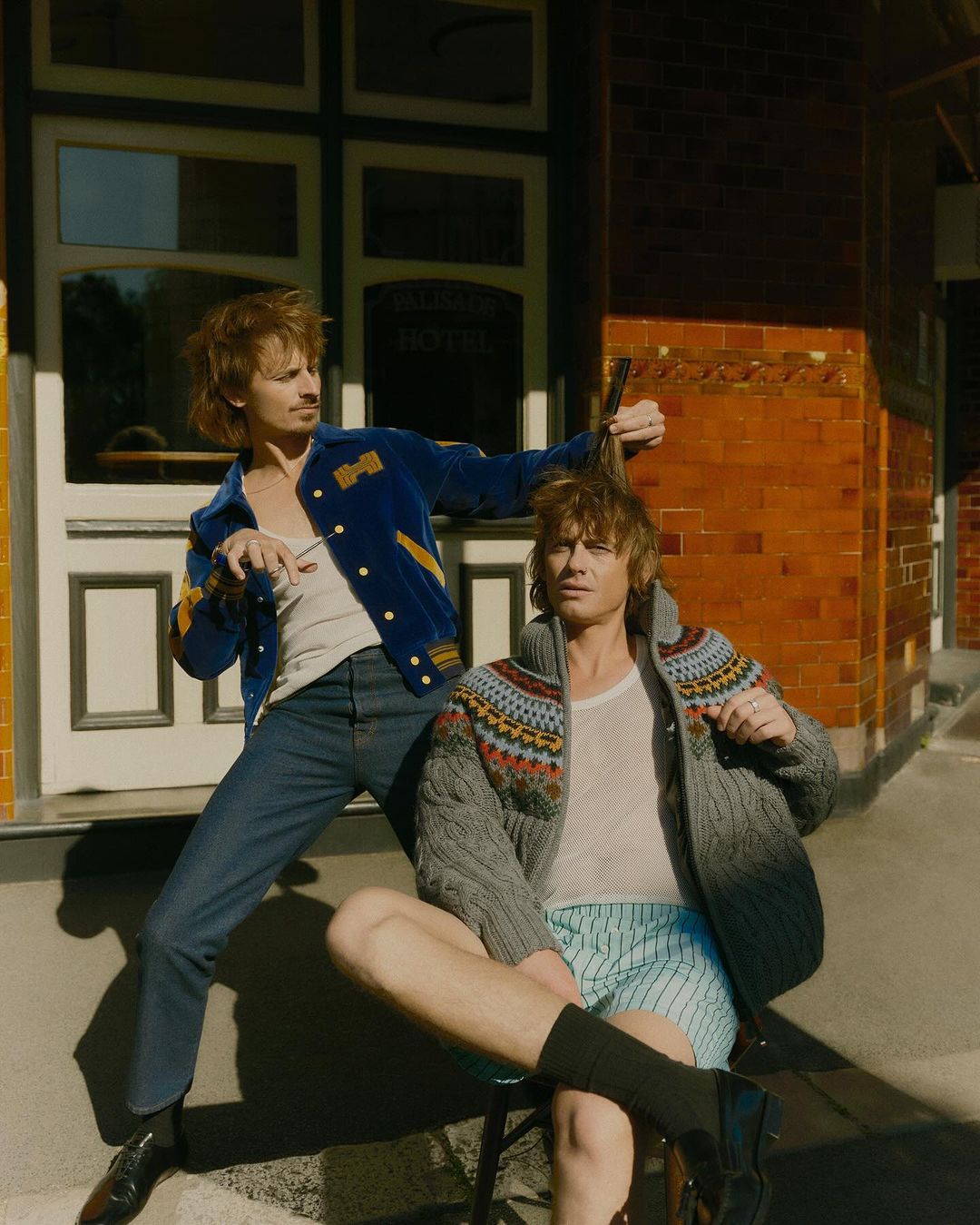 Two young men in a casual fashion shoot outside. One is cutting the other's hair with scissors and a comb in front of a brick building.