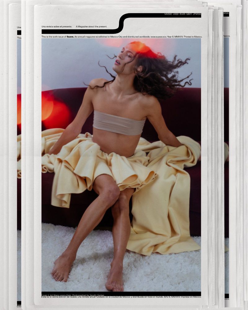 Fashion editorial photo of a slender adult with long hair wearing a wrap top and skirt, sitting on a couch on a magazine cover.