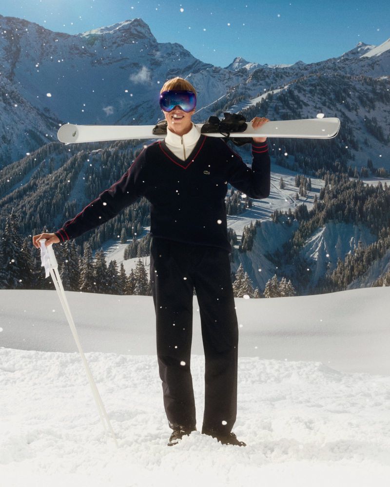 Man in ski goggles carrying skis and poles in snowy mountain landscape during winter. Wearing navy sweater and dark pants.