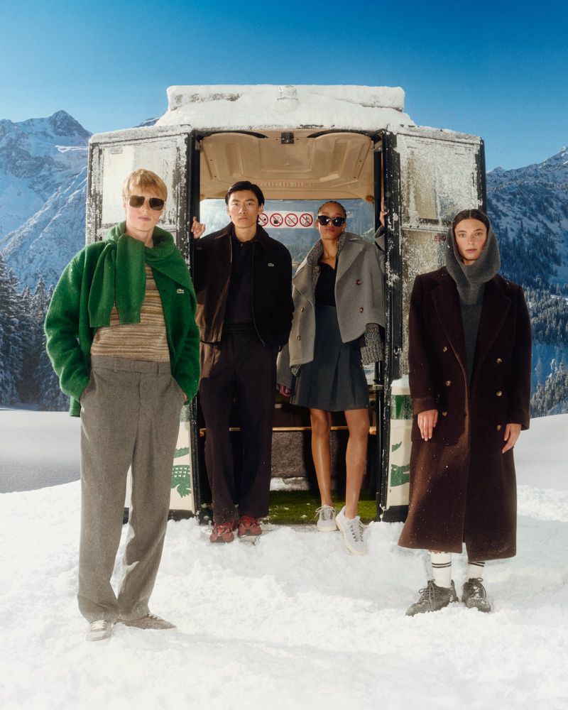 Group of four diverse adults posing in snow at a Lacoste-branded shuttle car wearing warm winter clothing in a snowy mountain landscape.