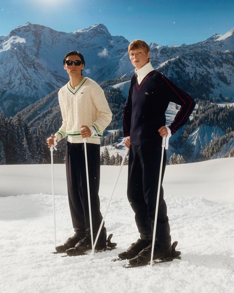 Two men in sweaters and ski attire pose on snowy mountain, trees and mountains visible in the background on a sunny day.
