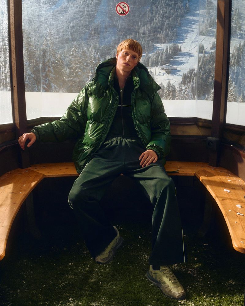Man in green puffer jacket sits in ski lift with snowy mountain view, winter fashion shoot.