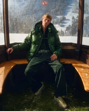 Man in green puffer jacket sits in ski lift with snowy mountain view, winter fashion shoot.