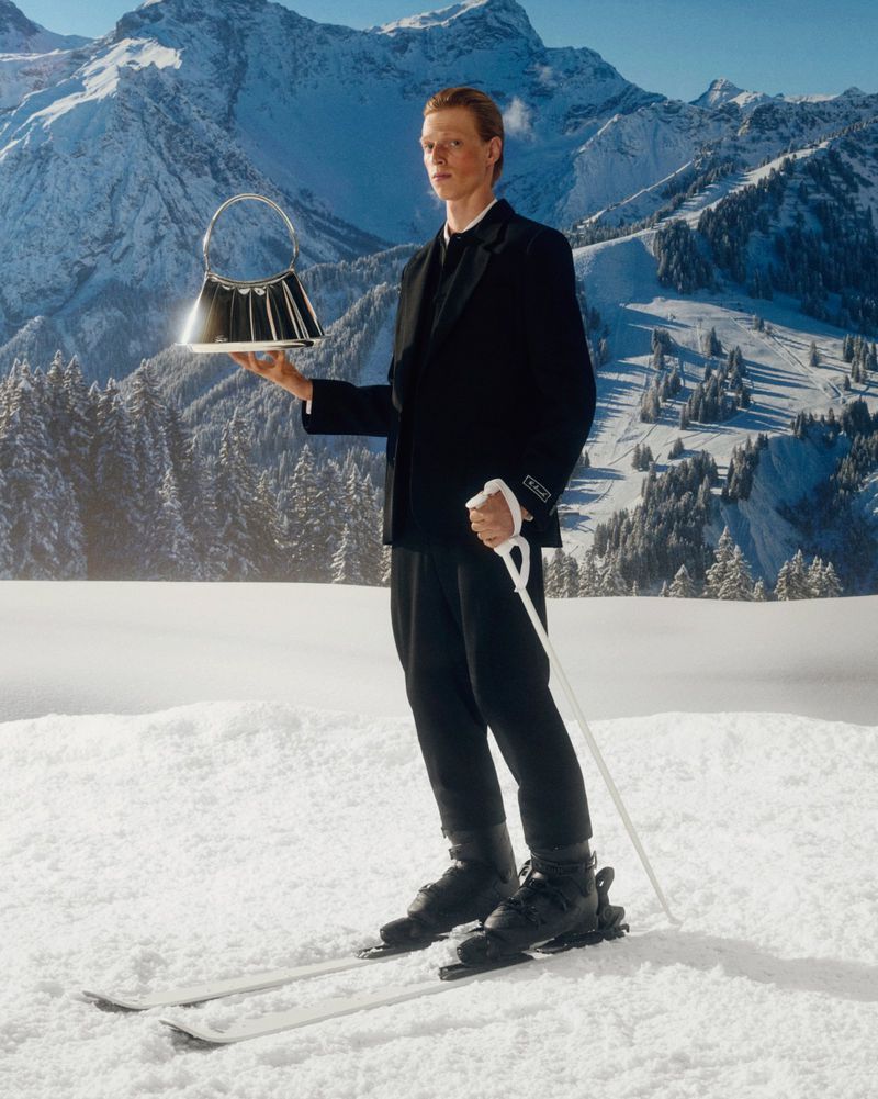Male model in ski boots, snow, holding purse against snow-capped mountains. Fashion, lifestyle shoot with dramatic alpine backdrop.