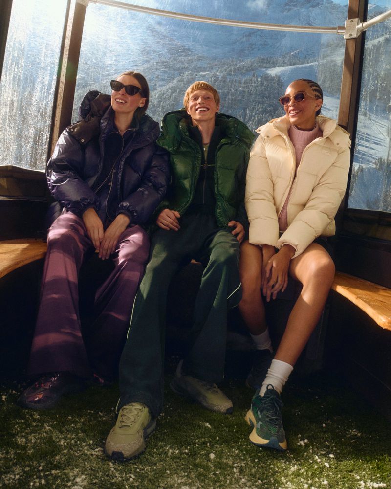 Three young adults, two women and one man, sit inside a ski gondola wearing winter jackets with a snowy mountain landscape in the background.