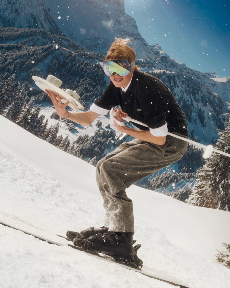 A person skis down a snowy slope with a mountain backdrop, wearing ski goggles and a Lacoste sweater, holding two plates of food.