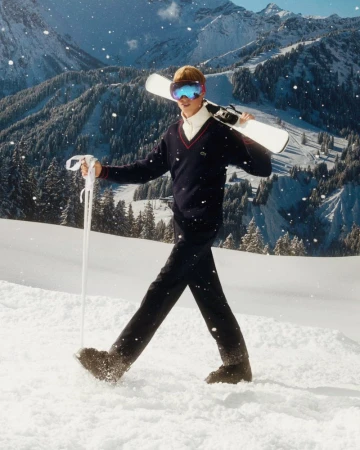 A man in ski goggles, carrying skis and ski poles, walks in a snowy mountain landscape with trees and mountain peaks.