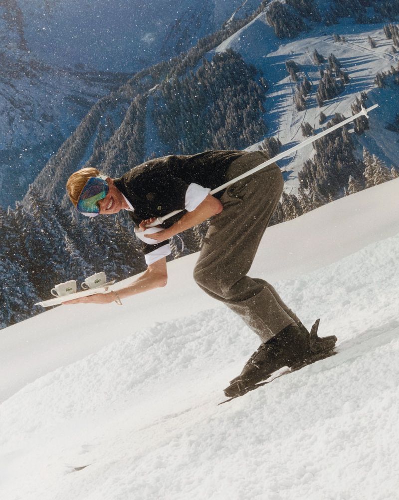 A woman skis down a snowy slope carrying a tray with two cups, with mountains and trees covered in snow in the background.