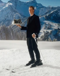 Model in a black ski outfit holding a shiny silver handbag on a snowy mountain landscape