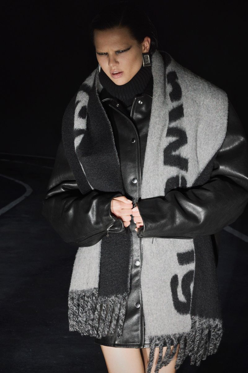 Woman in a black leather jacket and gray scarf poses in an outdoor setting with dark lighting for a fashion shoot.