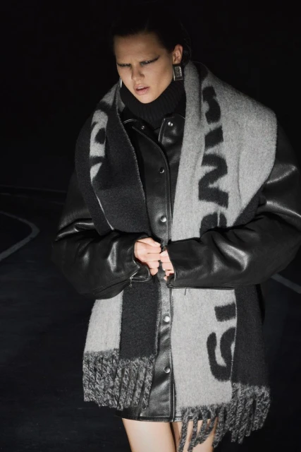 Woman in a black leather jacket and gray scarf poses in an outdoor setting with dark lighting for a fashion shoot.