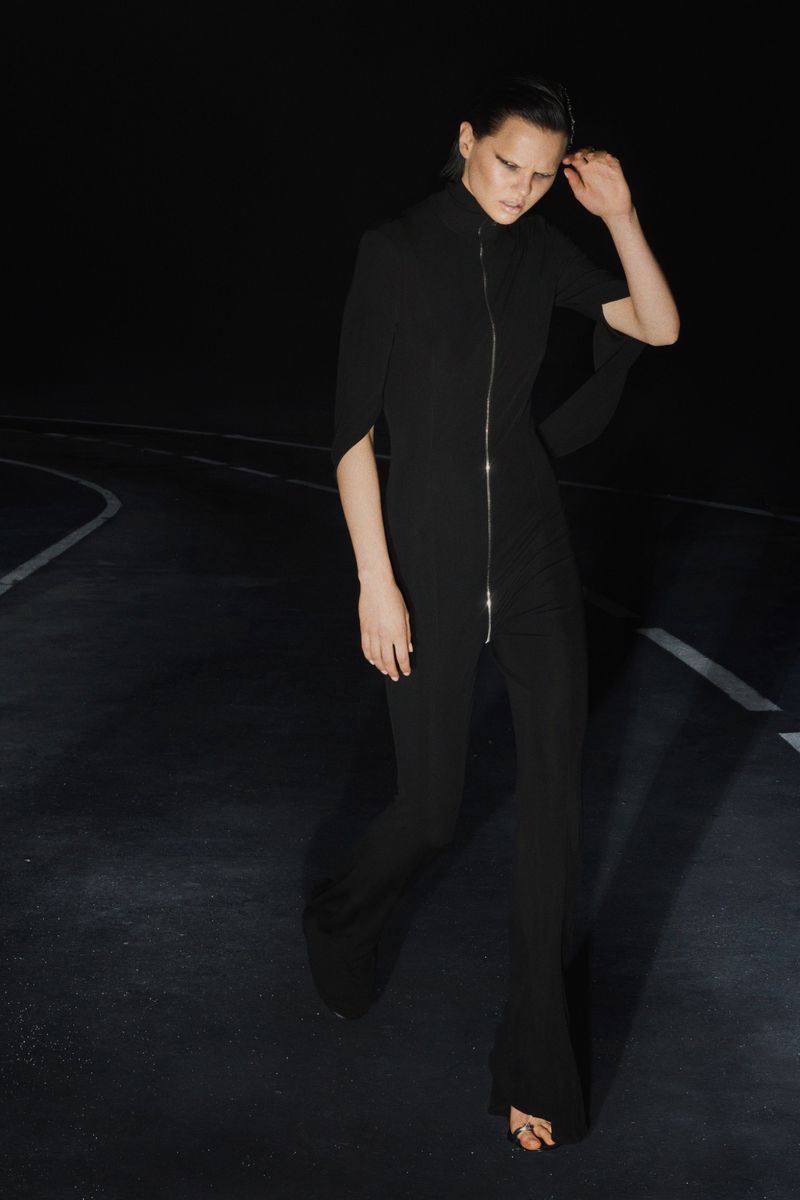 An adult female fashion model poses in a black jumpsuit on a dark street under dim lighting, showcasing the garment's design.