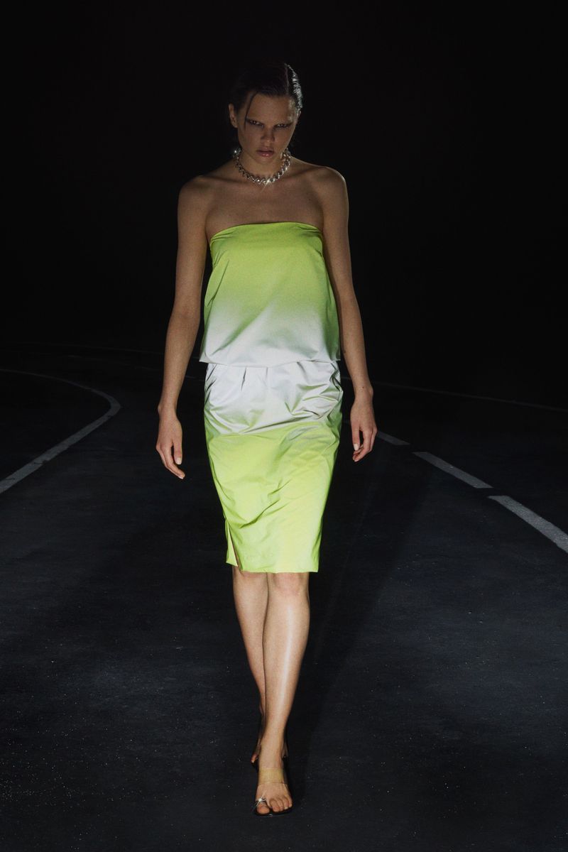 A woman models a strapless green dress on a dark runway, looking down as she walks barefoot towards the viewer in an editorial shot.