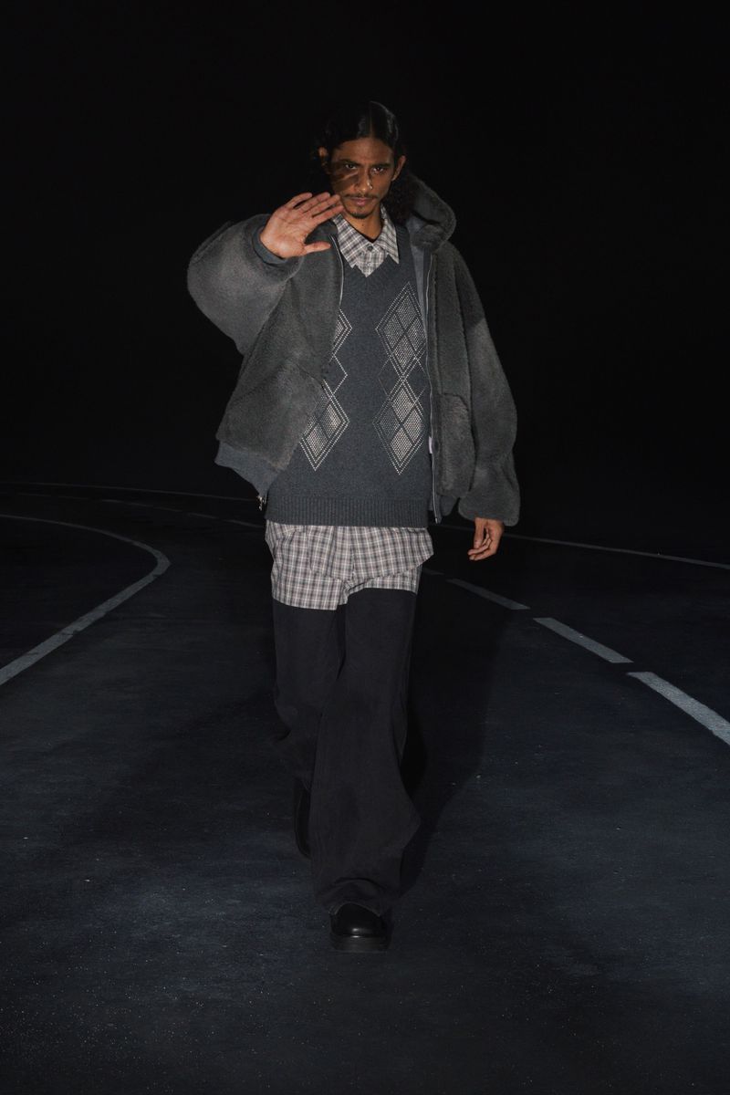 Adult male poses on dark road wearing a grey hooded jacket, patterned shirt, sweater vest, and black pants in a fashion editorial.