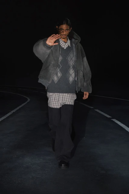 Adult male poses on dark road wearing a grey hooded jacket, patterned shirt, sweater vest, and black pants in a fashion editorial.